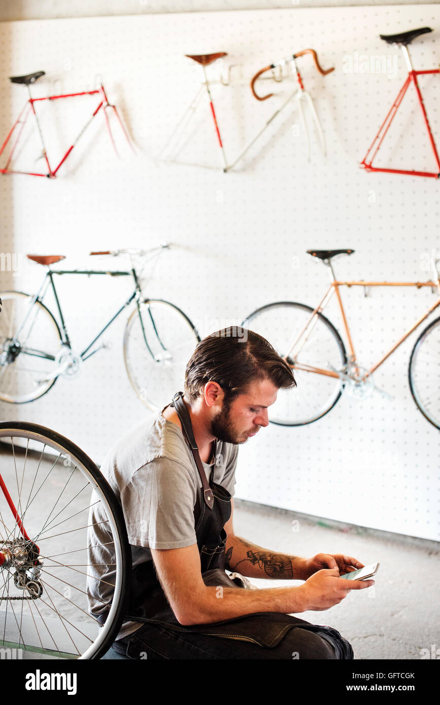 A man working in a bicycle repair shop sitting using his smart phone Stock Photo