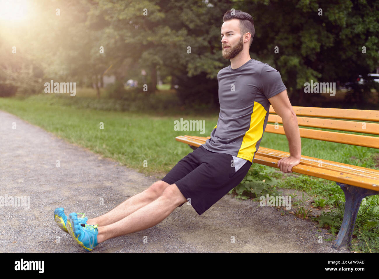 Young man exercising using a wooden park bench raising himself on his arms to tone his muscles, side view with copy space in a h Stock Photo