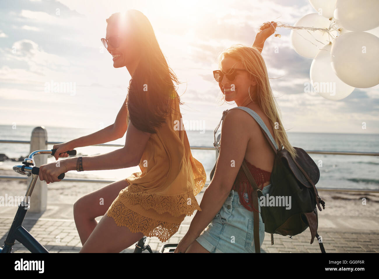 Happy young women riding a bicycle together with balloons. Best friends having fun on a cycle by the sea. Stock Photo