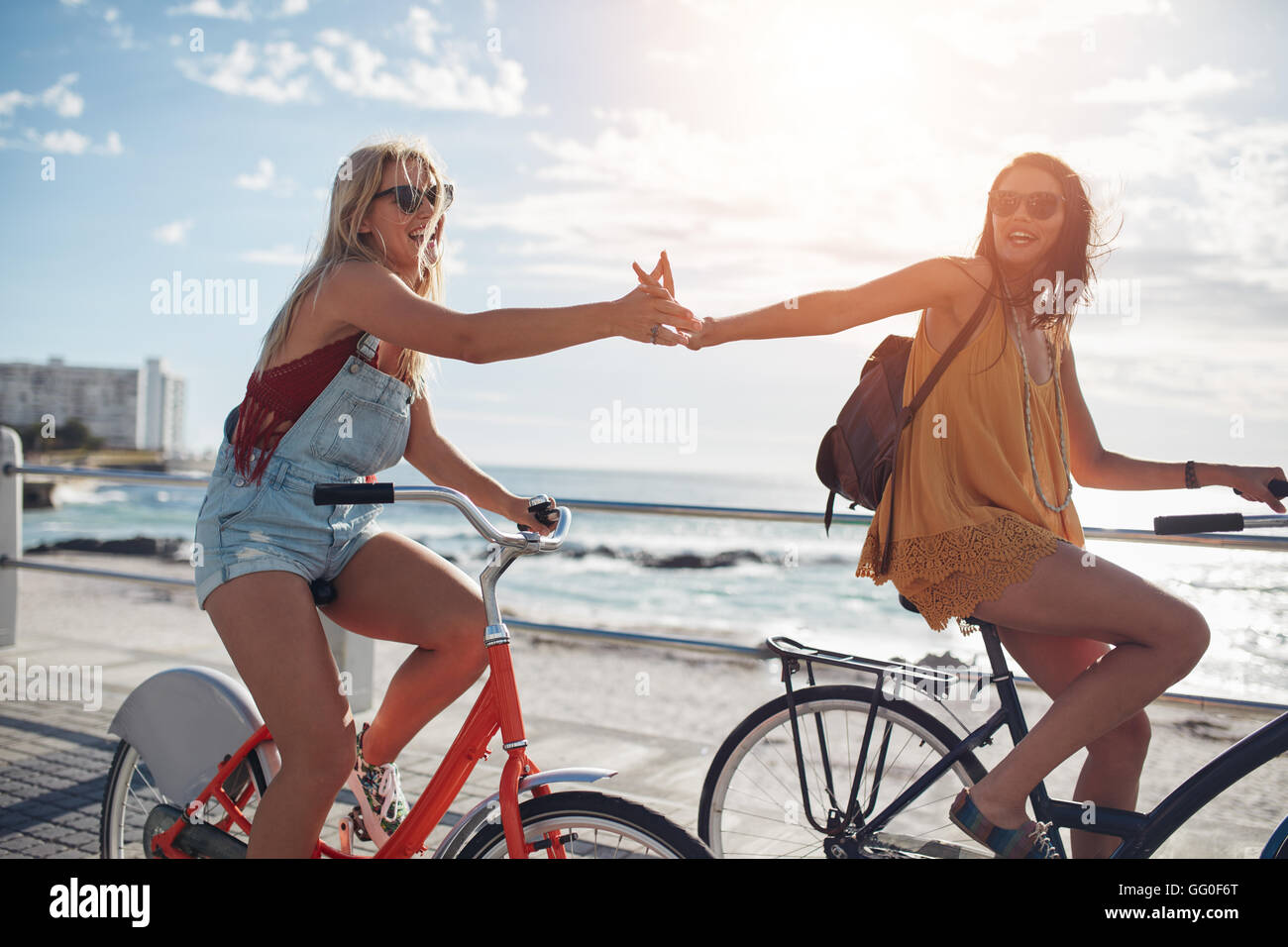 Female friends giving high five and riding bicycles along a seaside promenade on a summer day. Cheerful young women cycling outd Stock Photo