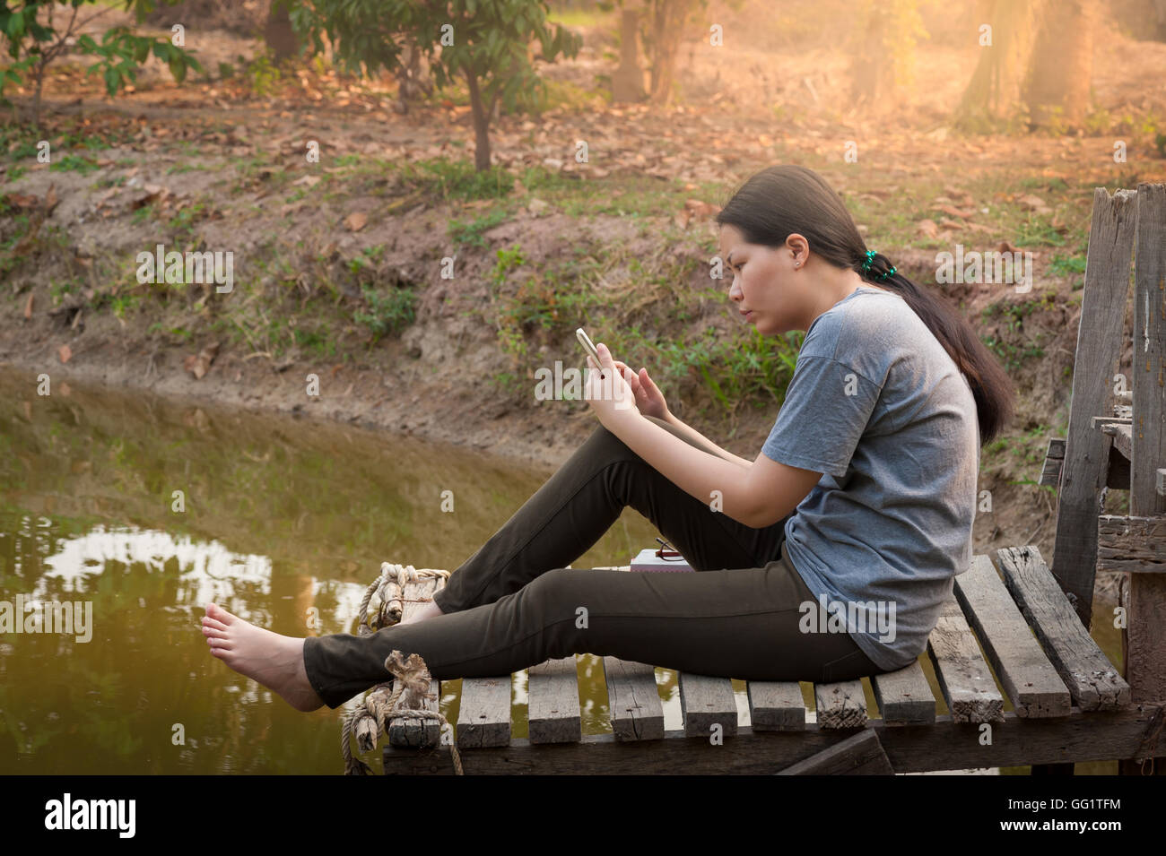 Weekend morning lifestyle. Young woman using her mobile phone seriously while sitting outdoor beside river in morning time. Stock Photo