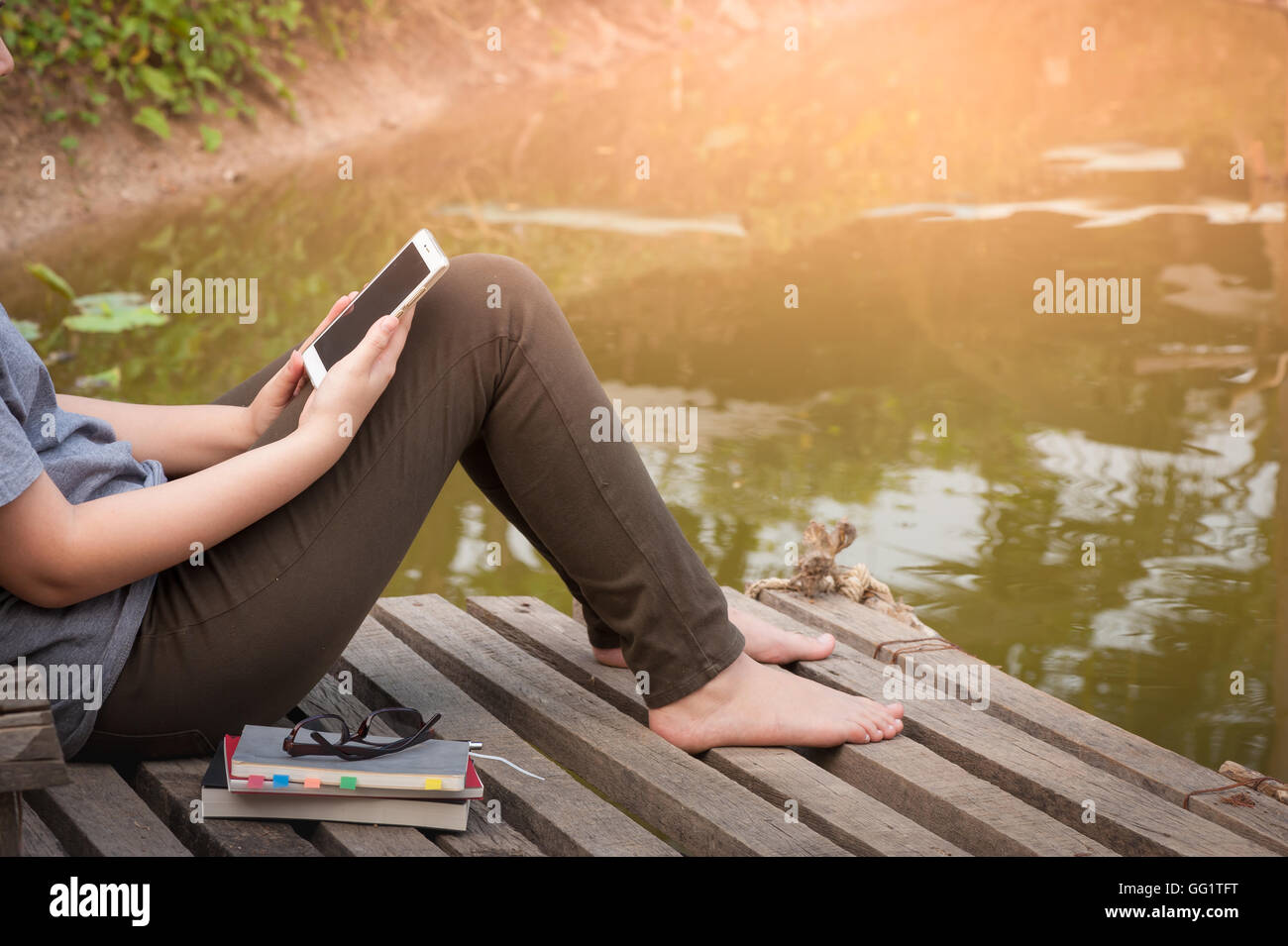 Weekend morning lifestyle. Young woman using her mobile phone seriously while sitting outdoor beside river with notebooks, pen, Stock Photo