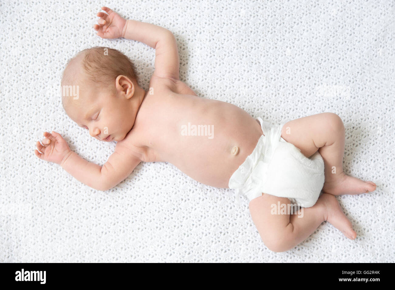 Portrait of young funny newborn babe napping on white knitted blanket with his arms up. Cute new born child sleeping. Healthy Stock Photo