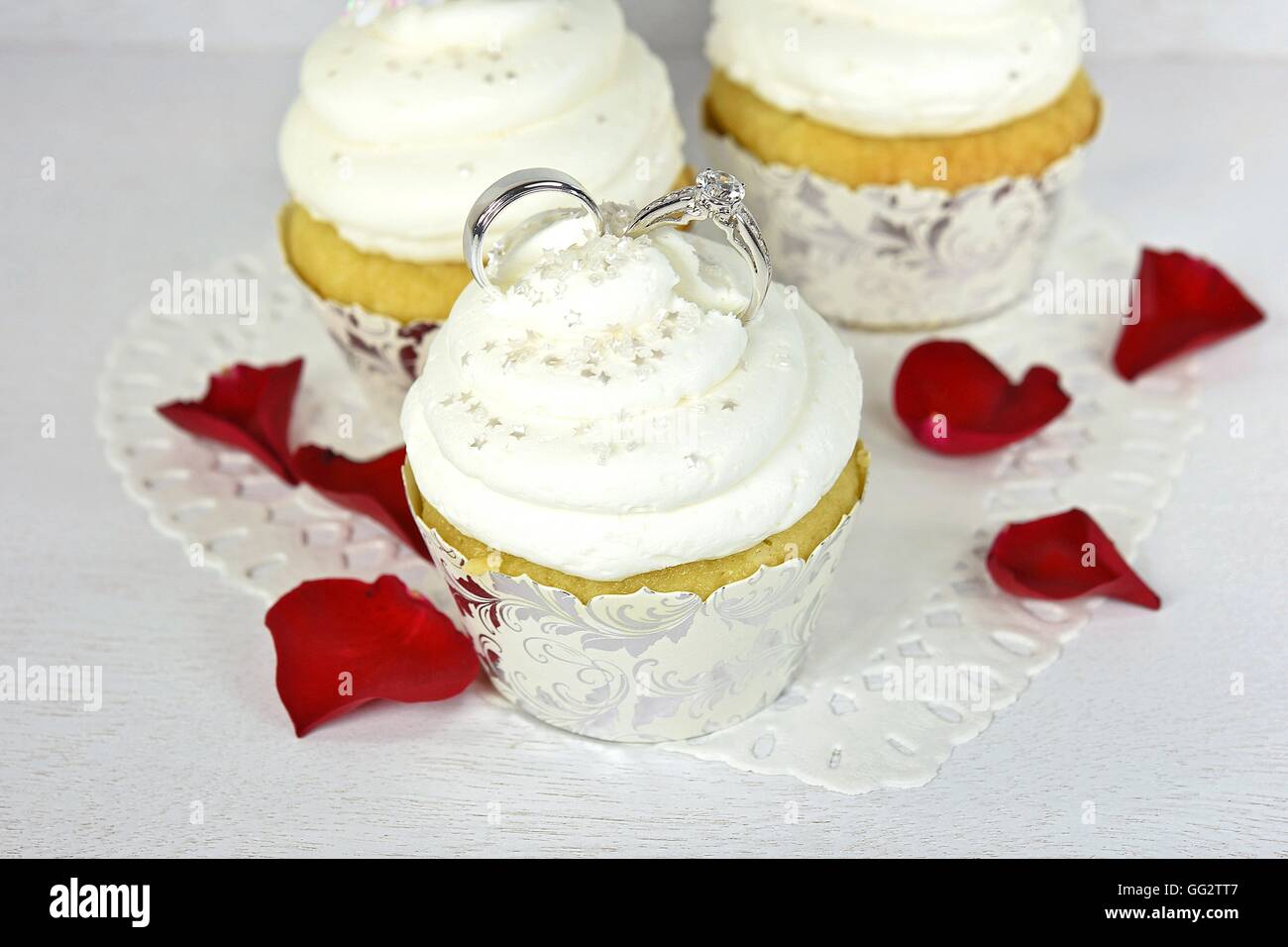 Wedding rings in cupcake icing with star sprinkles and red rose petals on paper heart doily. Stock Photo