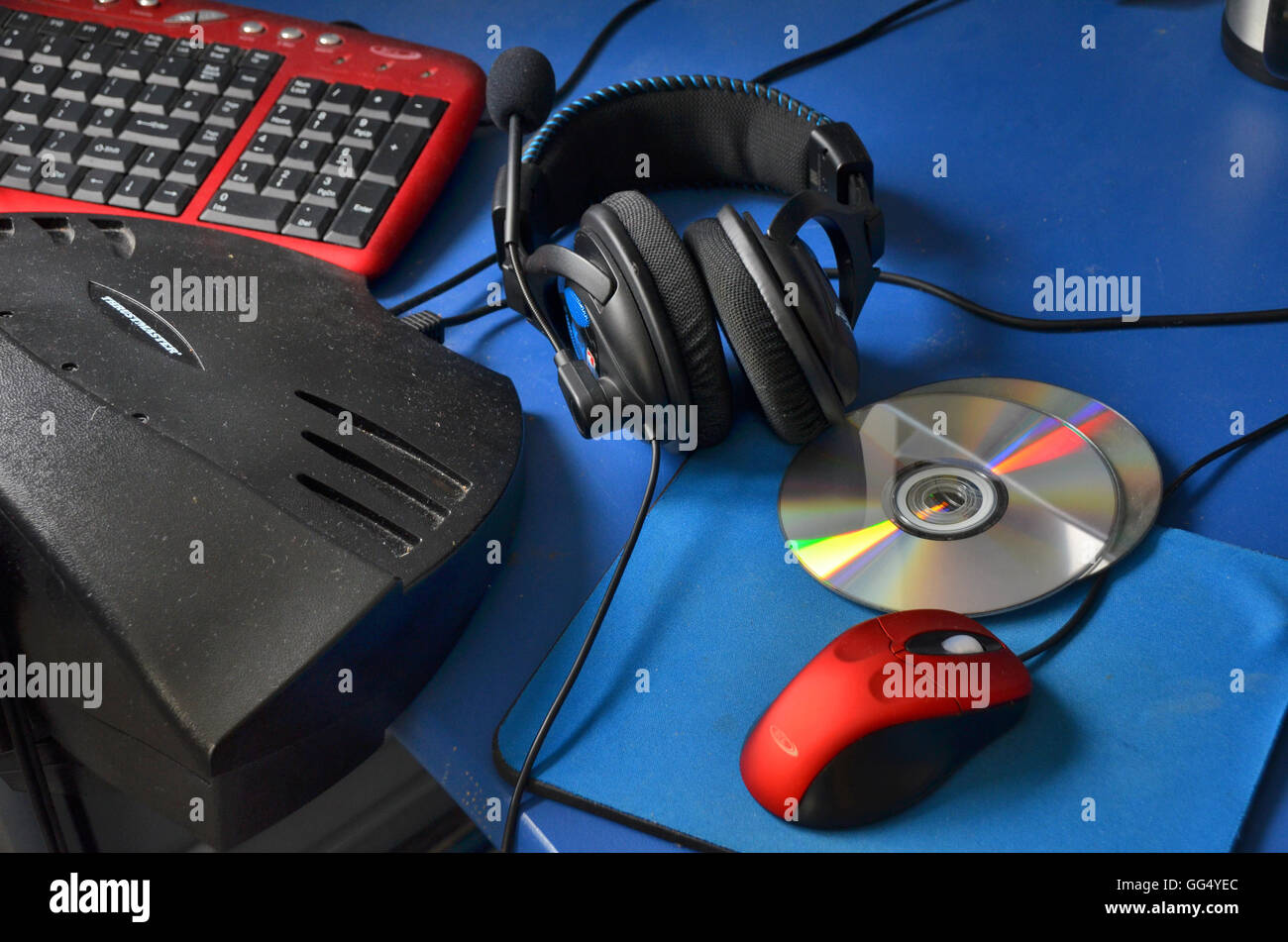 Computer Headphones, DVDs, red scroll mouse and mat and keyboard sit on a blue topped office desk. Stock Photo