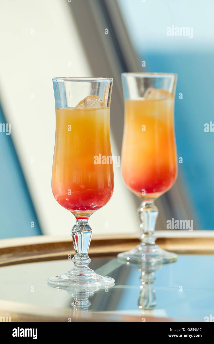 Two glasses containing Tequila Sunrise orange cocktail drink with ice on a glass table top beside a window. Stock Photo