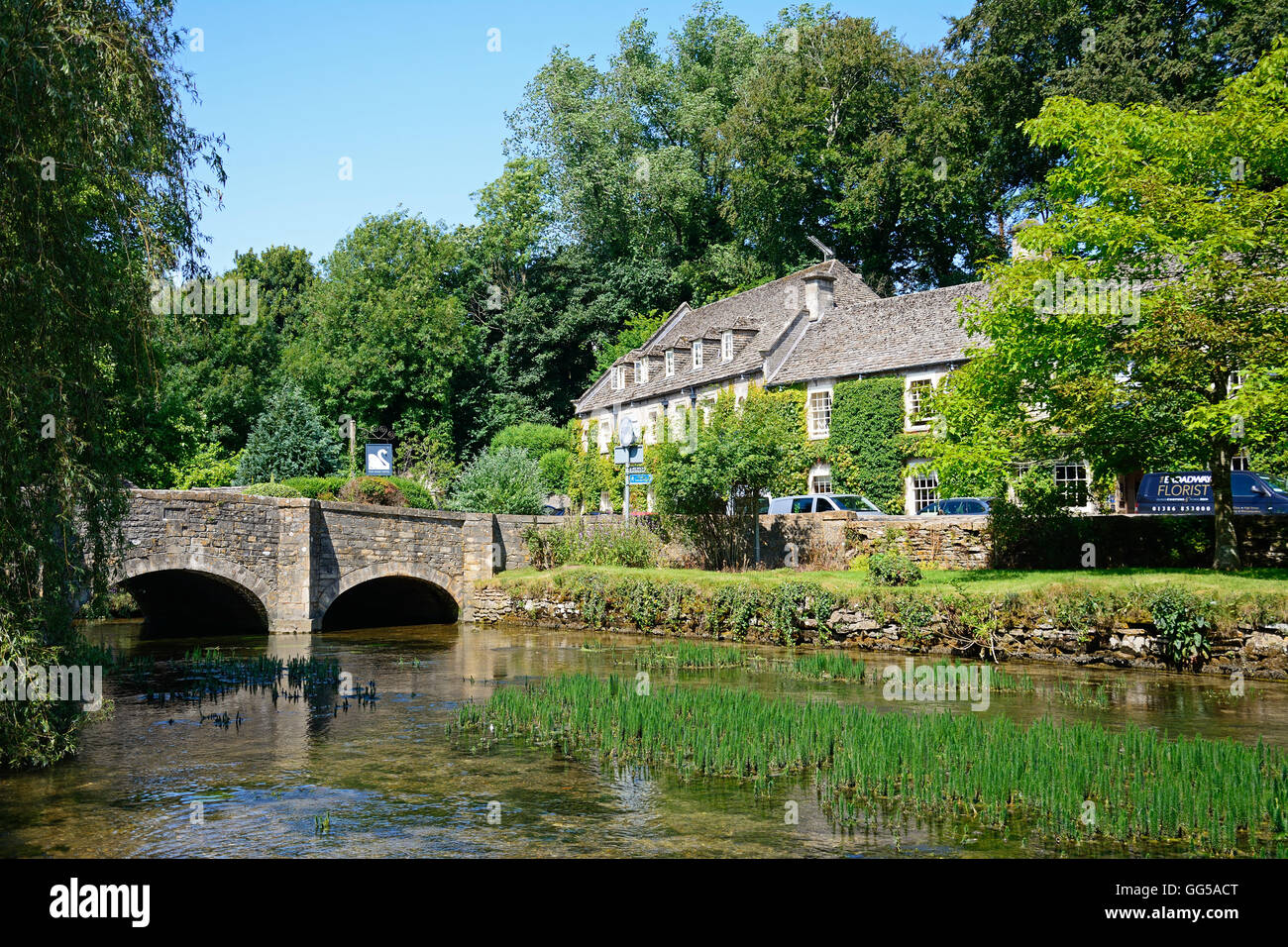 View along the River Coln towards the stone bridge with The Swan Hotel to the rear, Bibury, Cotswolds, Gloucestershire, England, Stock Photo