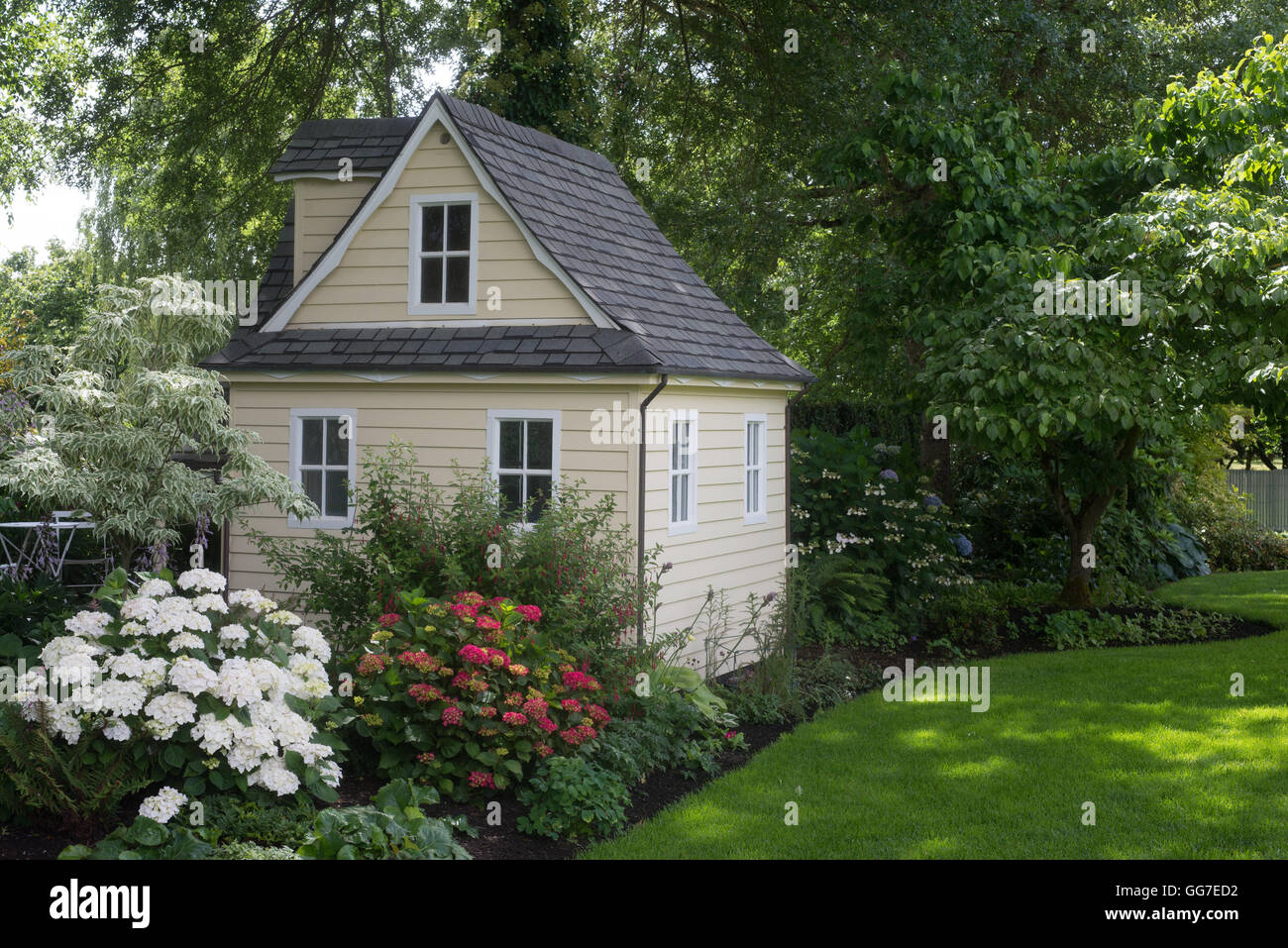 A charming playhouse cottage sits at the edge of a shaded perennial garden. Stock Photo