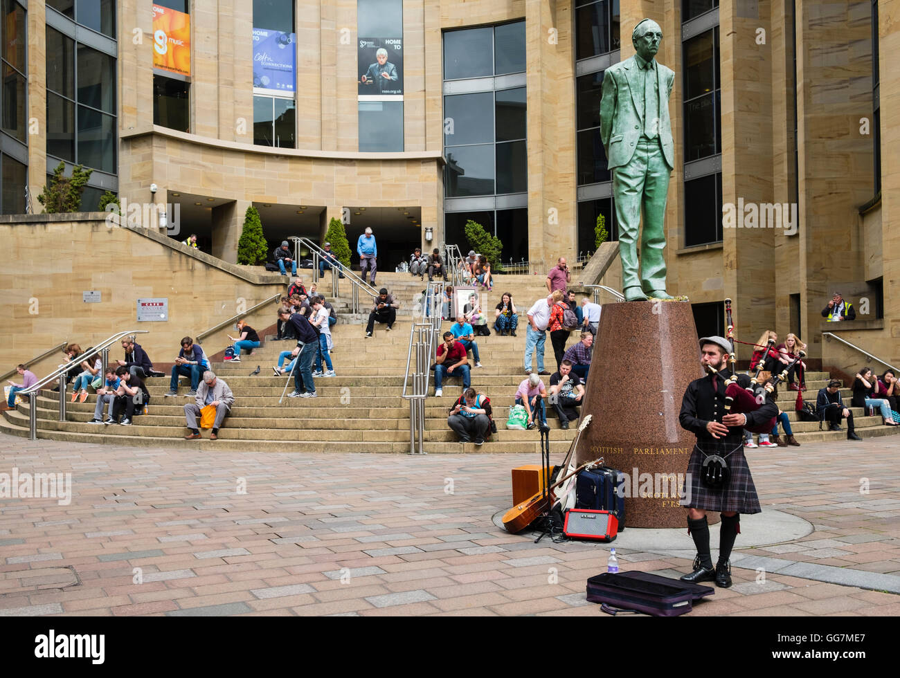 People sitting outside in the sun on steps of Glasgow Royal Concert Hall with busker playing bagpipes in Glasgow, Scotland, unit Stock Photo