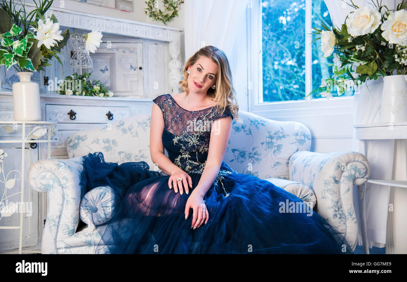Bridesmaid in blue dress in her boudoir surrounded by white flowers sitting on a sofa. Stock Photo