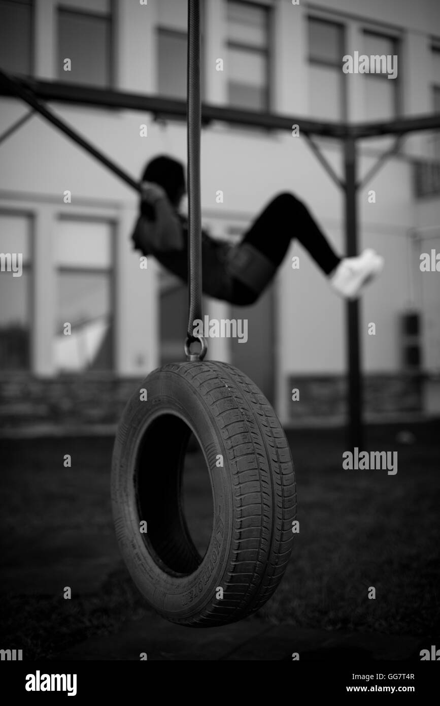 Old used car tyre hanging on a playground and a girl swinging on a swing in the background Stock Photo
