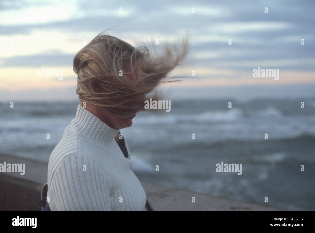 Woman's hair blowing in wind in front of sea waves and cloudy sky at sunset Stock Photo