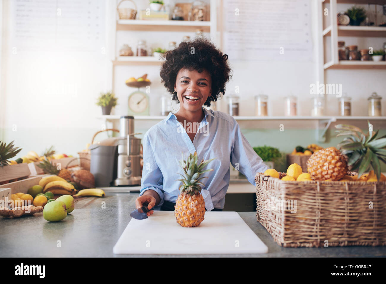 Portrait of cheerful young african woman standing behind bar counter with a pineapple on chopping board. Female working at juice Stock Photo