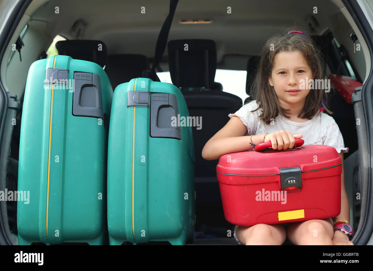 beautiful little girl with brown hair long loads the suitcase in the trunk of the car already full of suitcases before leaving f Stock Photo