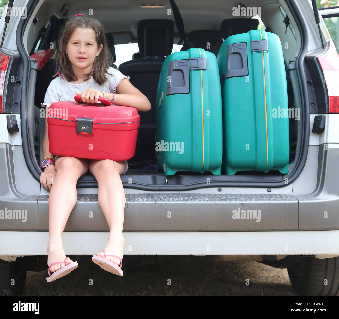 beautiful little girl with brown hair long loads the suitcase in the trunk of the car already full of bagagge before leaving for Stock Photo