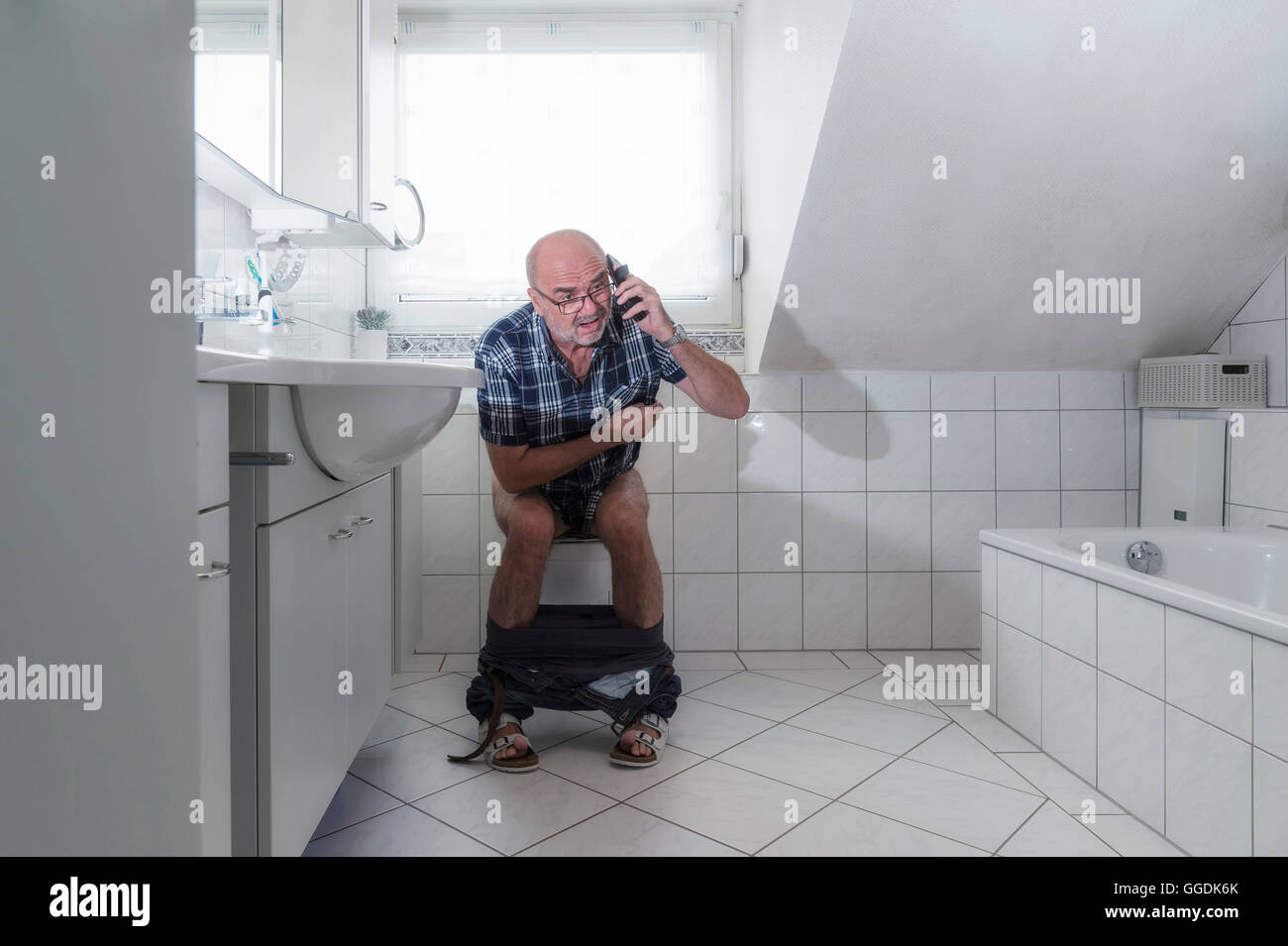 Senior man sitting on the toilet, talking into a smartphone, Germany Stock Photo