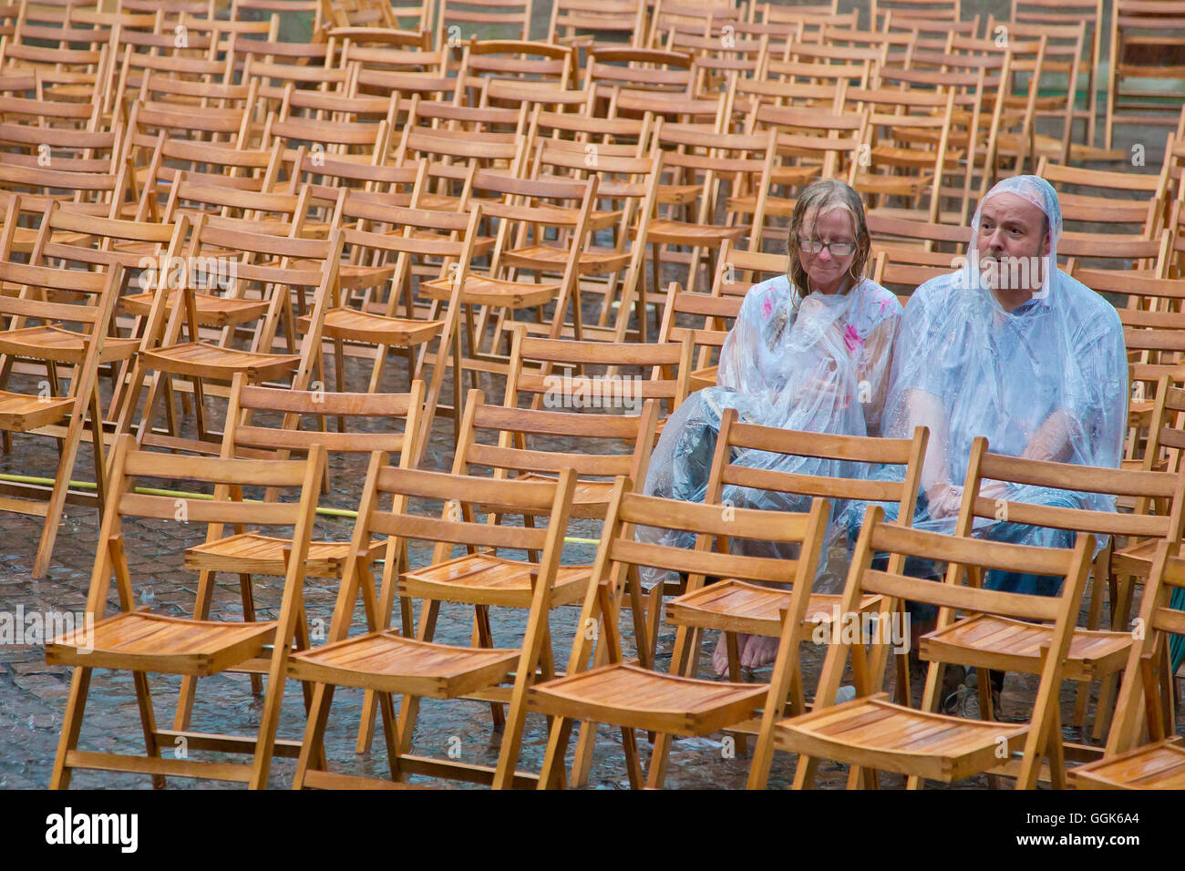 A couple wearing rain capes sitting on their own amidst wooden chairs in the pouring rain outside Friedrichstein Palace, Bad Wil Stock Photo