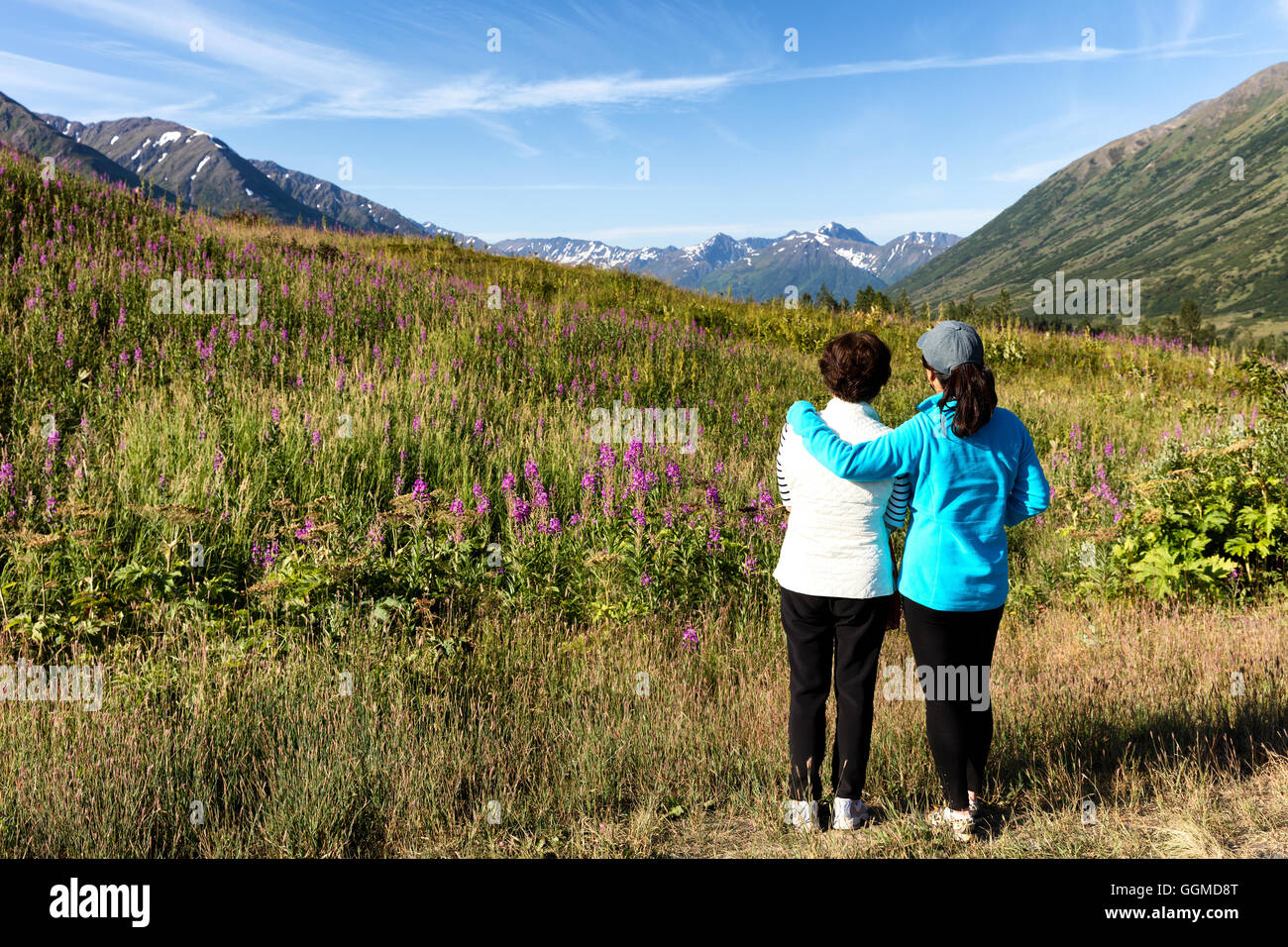 Mother and daughter, backs to camera, looking at wild flowers in field with mountains and sky in background. Selective focus on Stock Photo