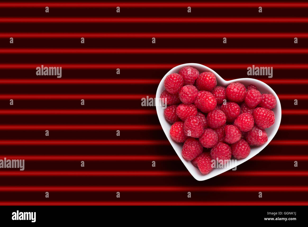 Raspberries in heart-shape bowl on red striped background Stock Photo