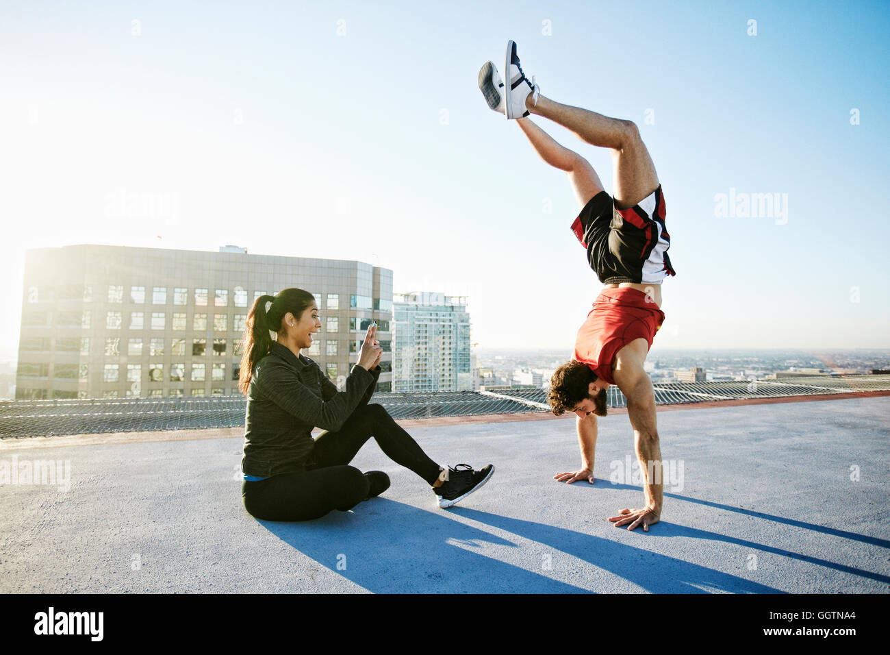 Woman photographing man doing handstand on urban rooftop Stock Photo