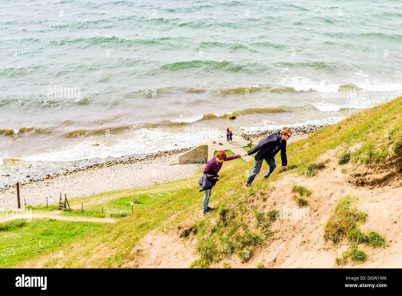 Kaseberga, Sweden - August 1, 2016: Young adult man helping a woman climb a steep sandy hill from the ocean below. Real life sit Stock Photo