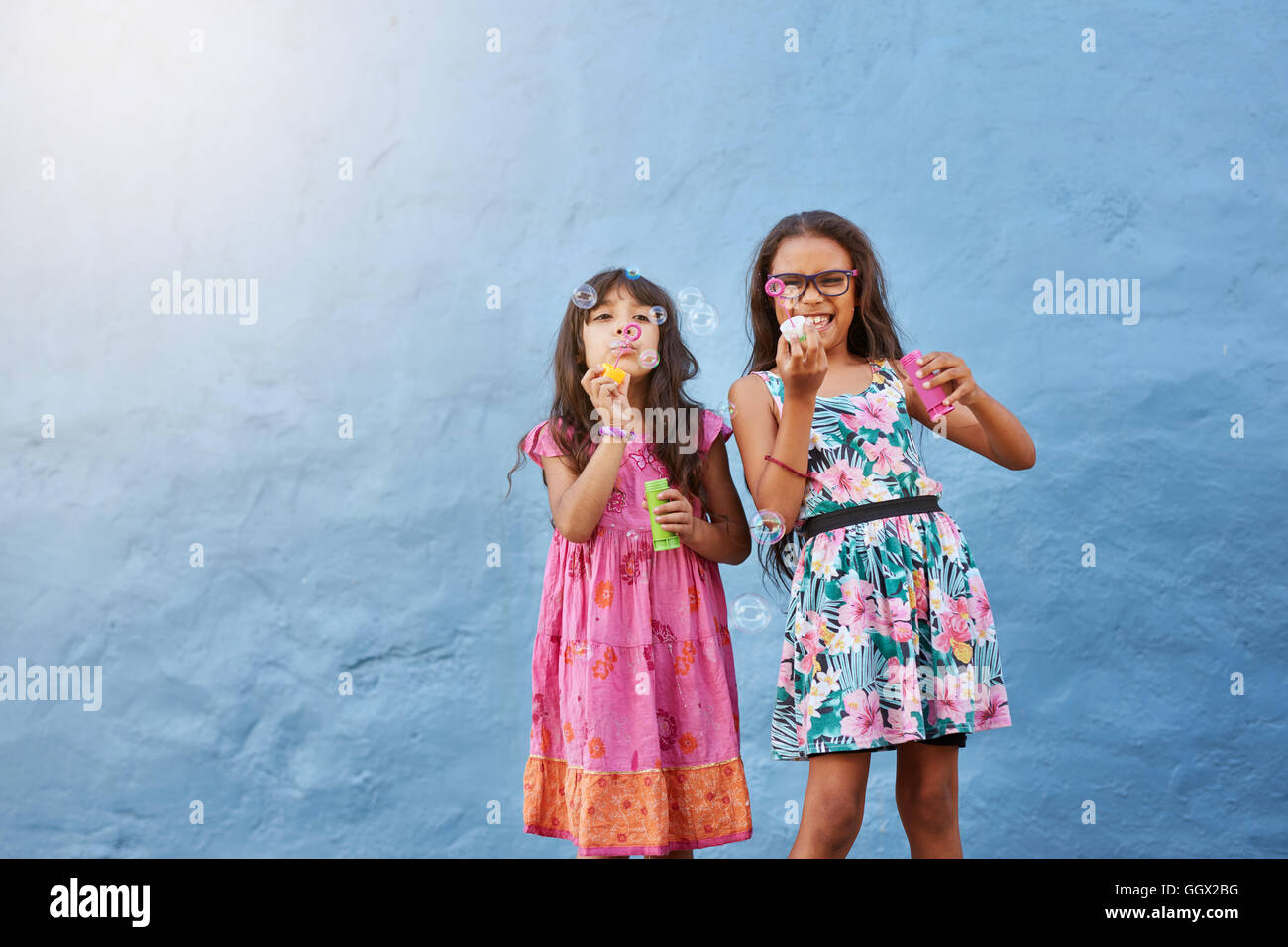 Portrait of cute little girls blowing soap bubbles. Two young girls playing against blue background. Stock Photo