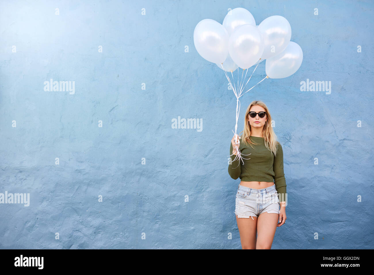 Horizontal shot of attractive female fashion model holding balloons against blue wall. Attractive young woman holding white ball Stock Photo