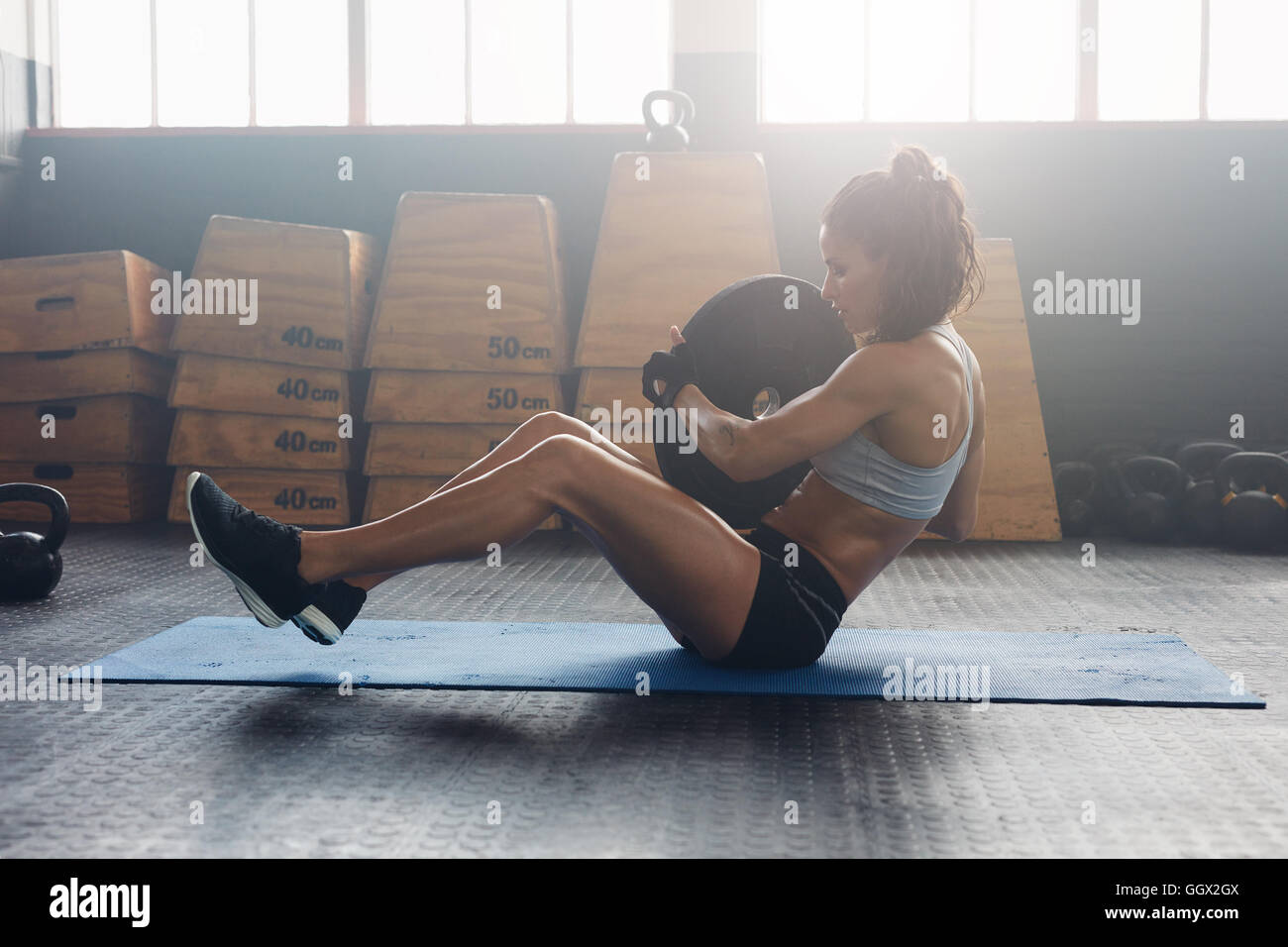 Side view shot of energetic woman doing abdominal exercise at fitness center. Woman athlete doing situps at the gym holding heav Stock Photo