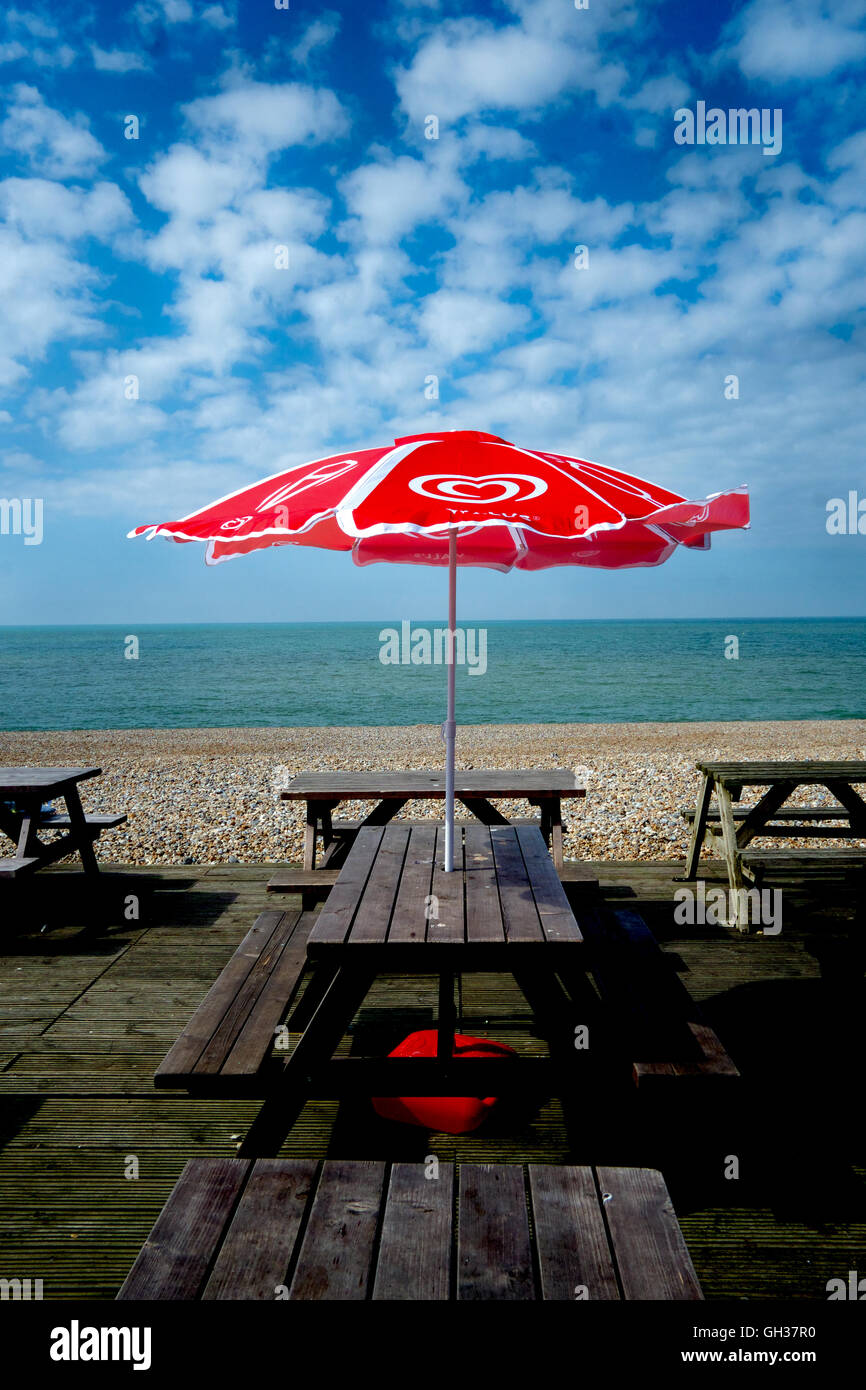 Red & white umbrella on wooden table and benchs in seaside cafe, sea and beach in background, blue sky with white clouds Stock Photo