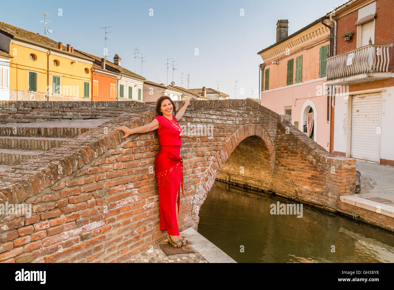 Mature Woman With Skin Marked By Wrinkles And Age Spots In Red Glass Necklace And Dress With Shawl On Hips Smiling While Leaning Against A Brick Wall Of An Old Bridge Of Mature Woman With Skin Marked By Wrinkles And Age Spots In Red Glass Necklace And Dress With Shawl On Hips Smiling While Leaning Against A Brick Wall Of An Old Bridge Of
