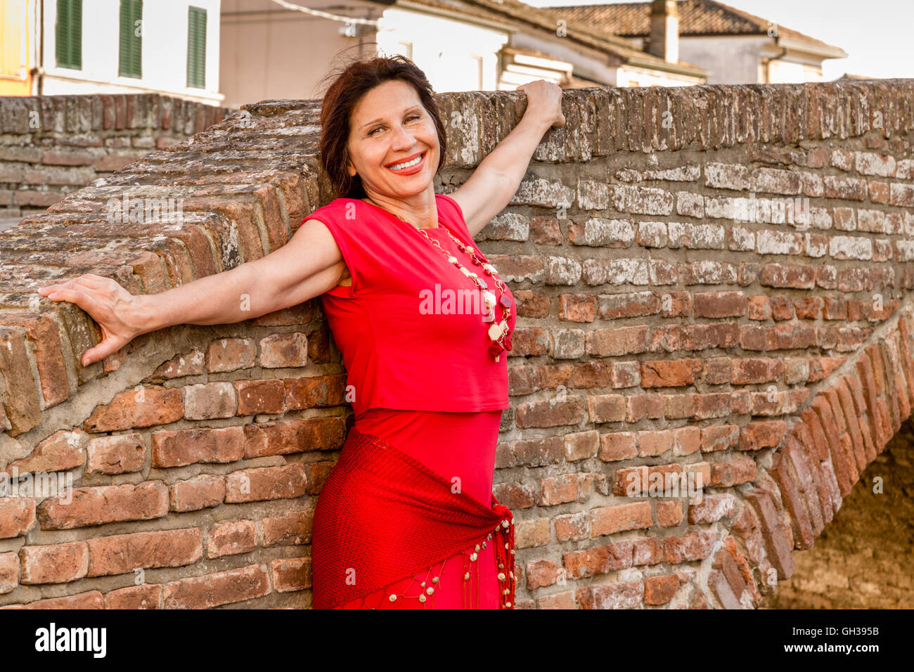 Mature Woman With Skin Marked By Wrinkles And Age Spots In Red Glass Necklace And Dress Smiling While Leaning Against A Brick Wall Of An Old Bridge Of A Typical Town In Mature Woman With Skin Marked By Wrinkles And Age Spots In Red Glass Necklace And Dress Smiling While Leaning Against A Brick Wall Of An Old Bridge Of A Typical Town In