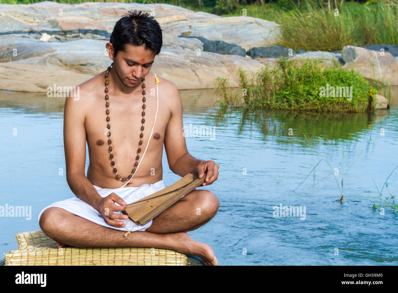 A young brahmin reads an ancient Hindu palm-leaf scripture on a river bank. Stock Photo