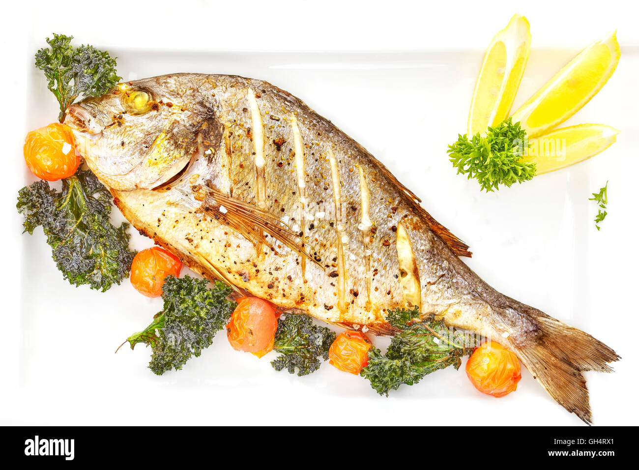 Roasted gilt head bream fish on a white plate with grilled tomatoes, kale and coarse grained salt. Stock Photo