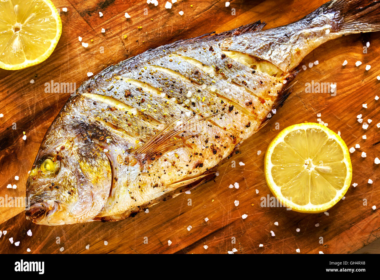 Roasted gilt head bream fish on a wooden table with lemons and coarse grained salt. Stock Photo