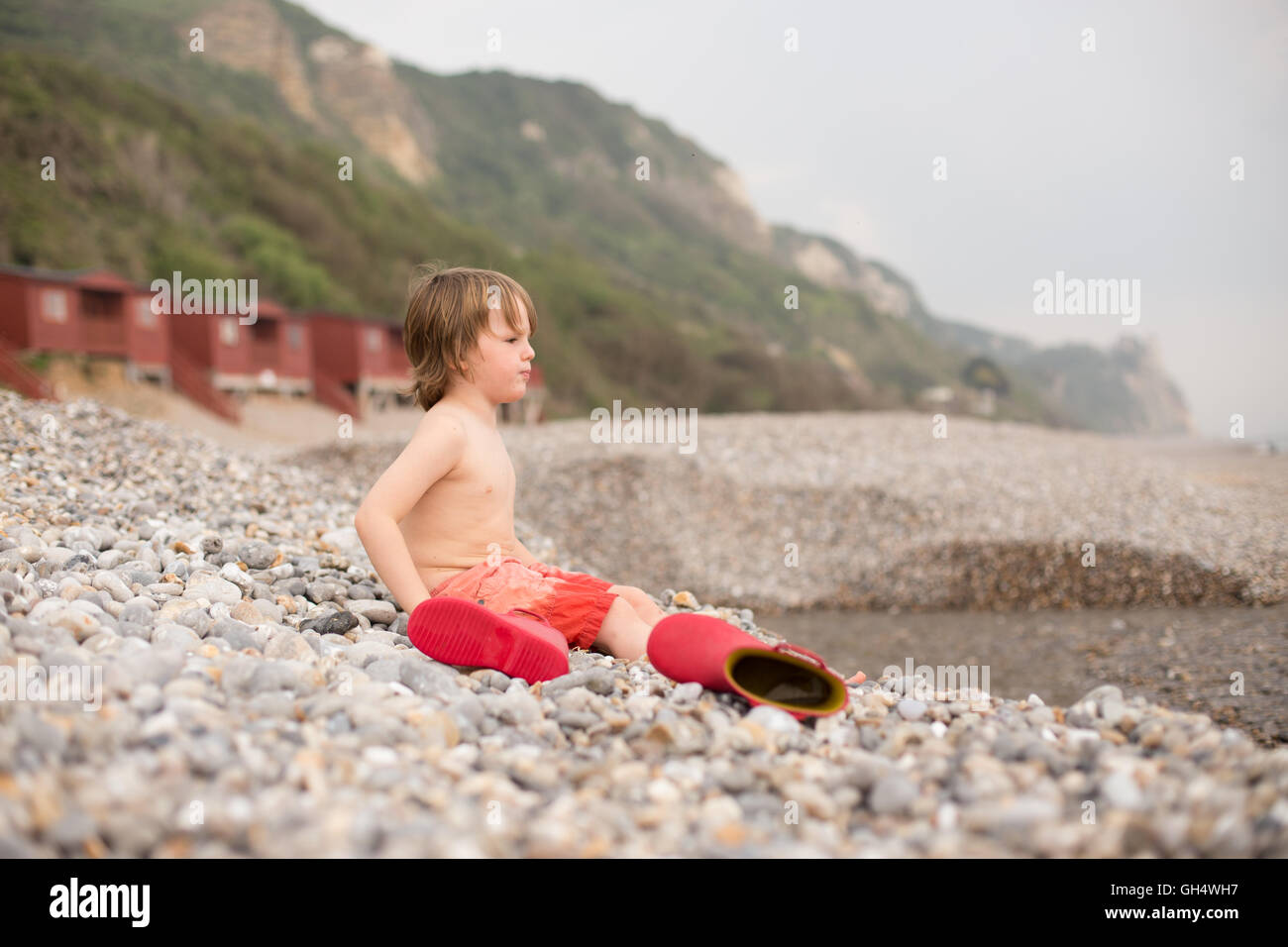 cross boy on pebble beach in red  wellies, beach huts houses in background and cliffs Stock Photo