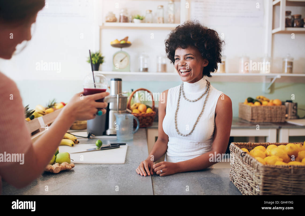 Portrait of smiling young african woman standing behind juice bar counter and talking with customer holding a glass of fresh jui Stock Photo