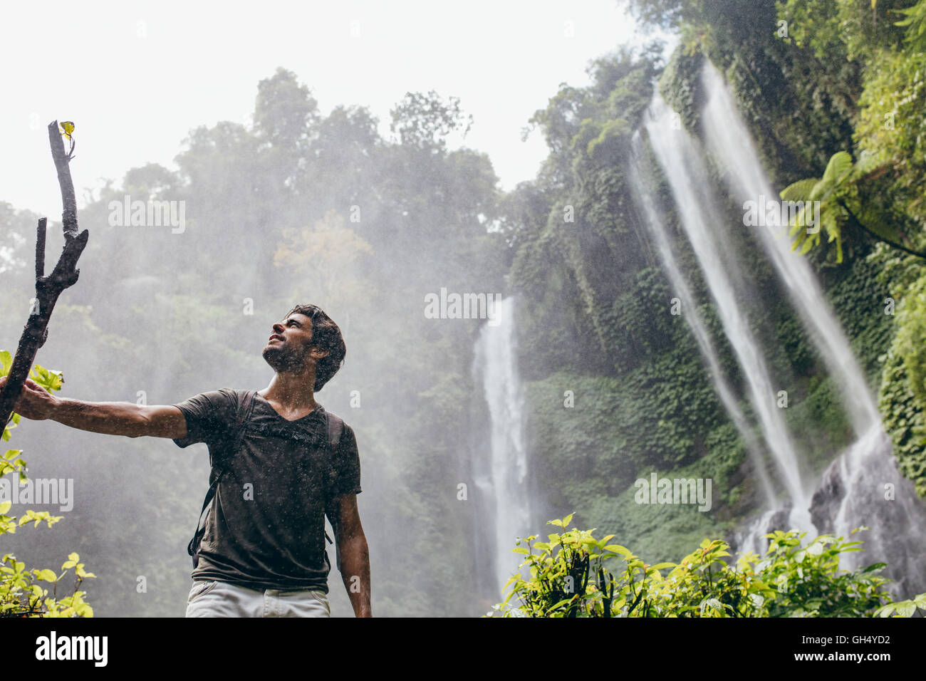Low angle shot of young man standing near a waterfall in forest and looking away. Male hiker enjoying in the nature with rain. Stock Photo