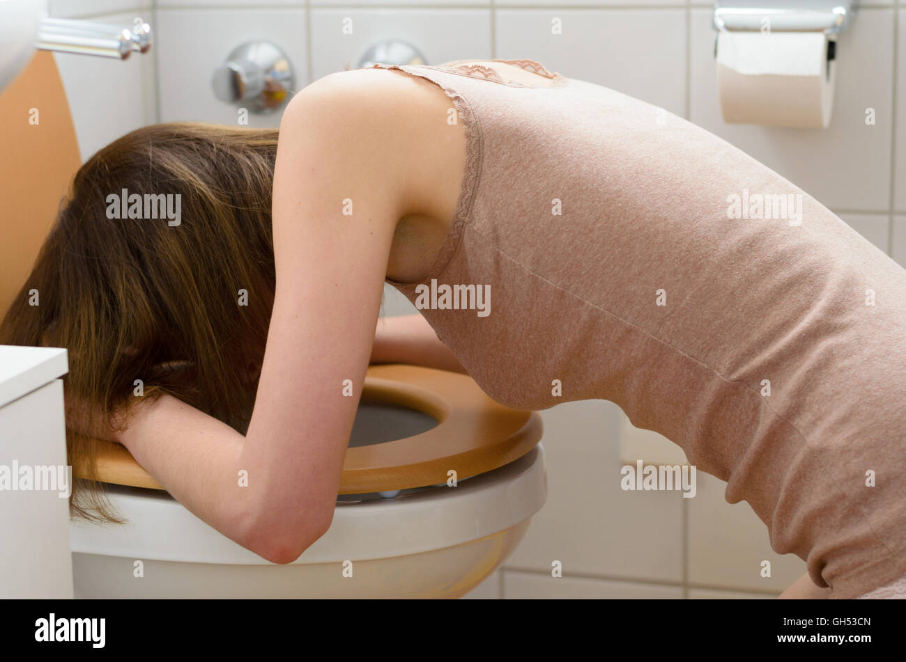 Side view on sick young woman wearing sleeveless shirt in leaning on open toilet seat at indoor bathroom Stock Photo