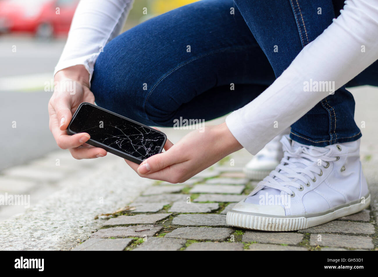 Young woman picking up her smashed mobile phone after dropping it on the cement paving of the sidewalk showing the shattered dis Stock Photo