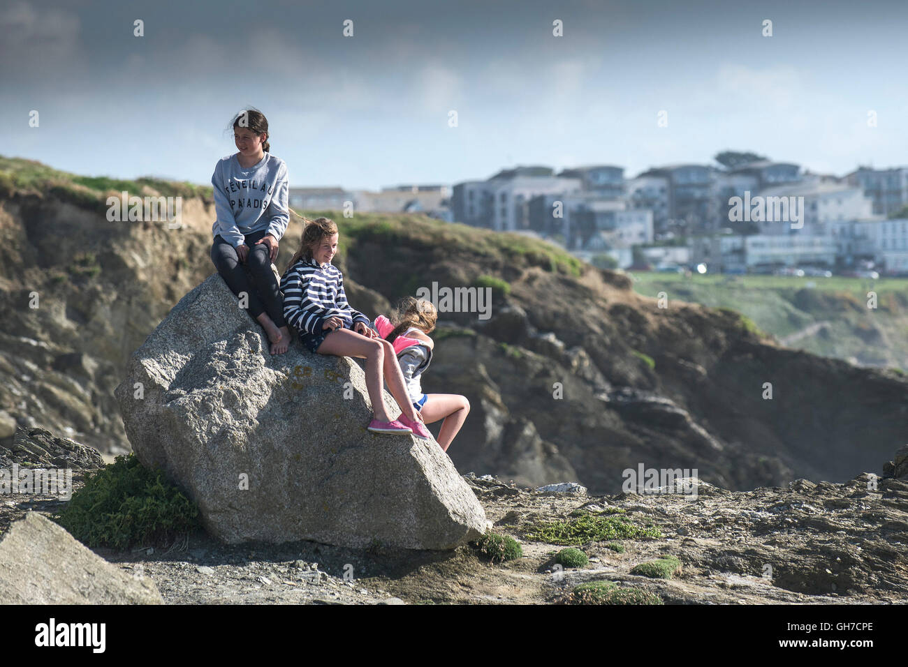 Three young girls sit on a rock on Towan headland in Newquay, Cornwall. Stock Photo