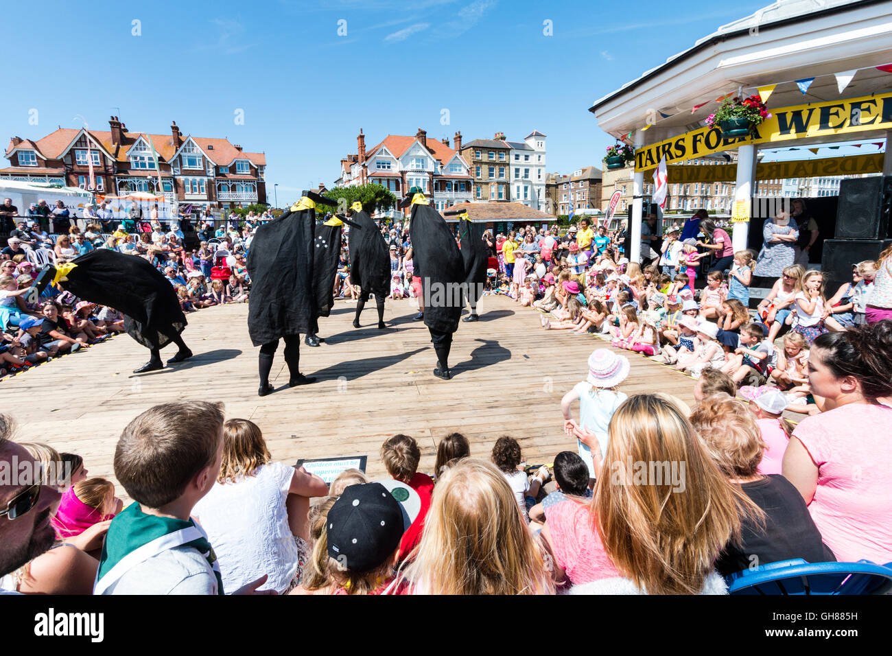 Popular Hobby Horse Club event for families and children at the Broadstairs folk week. Ossies, black bird type creatures each with a man inside, dancing on the deck by the bandstand, surrounding by audience of children and parents. Stock Photo