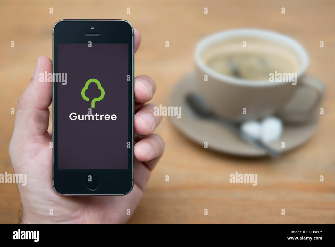 A man looks at his iPhone which displays the Gumtree logo, while sat with a cup of coffee (Editorial use only). Stock Photo