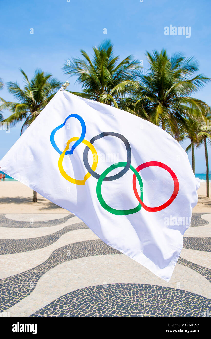 RIO DE JANEIRO - MARCH 10, 2016: Olympic flag waves in front of the iconic wave pattern of the sidewalk on Copacabana Beach. Stock Photo