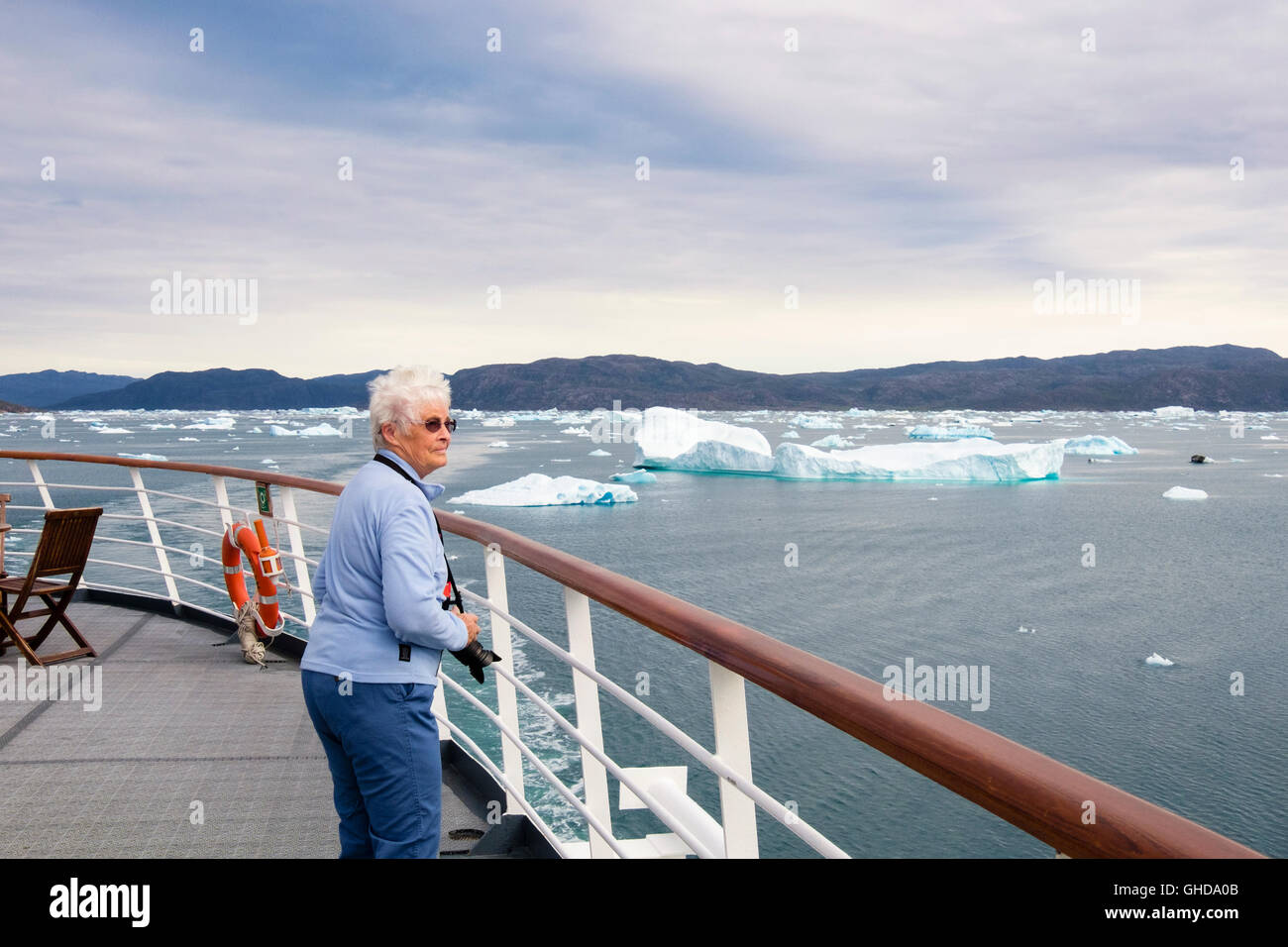Retired senior female tourist passenger on outside deck of a cruise ship sailing in Bredefjord fjord with icebergs in summer. Narsaq Greenland Stock Photo