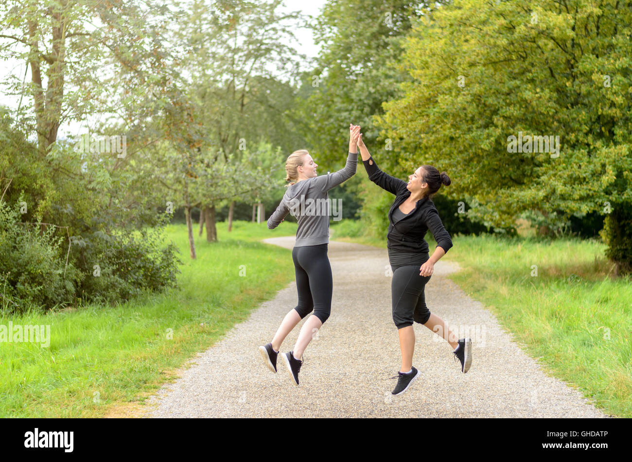 Pair of happy jumping women in workout clothes congratulating each other by slapping palms in mid air over running path Stock Photo