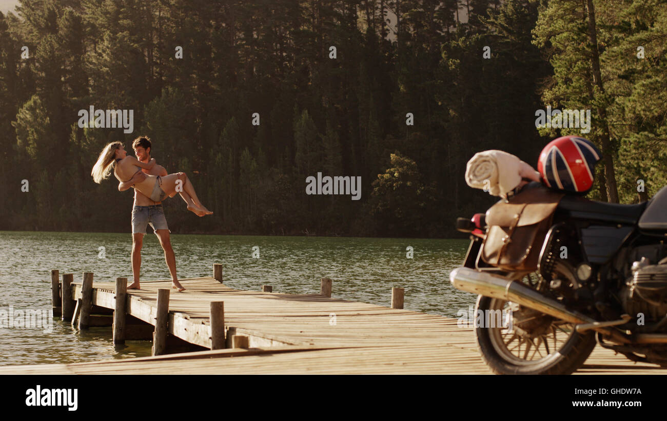 Young man carrying young woman on lakeside dock near motorcycle Stock Photo