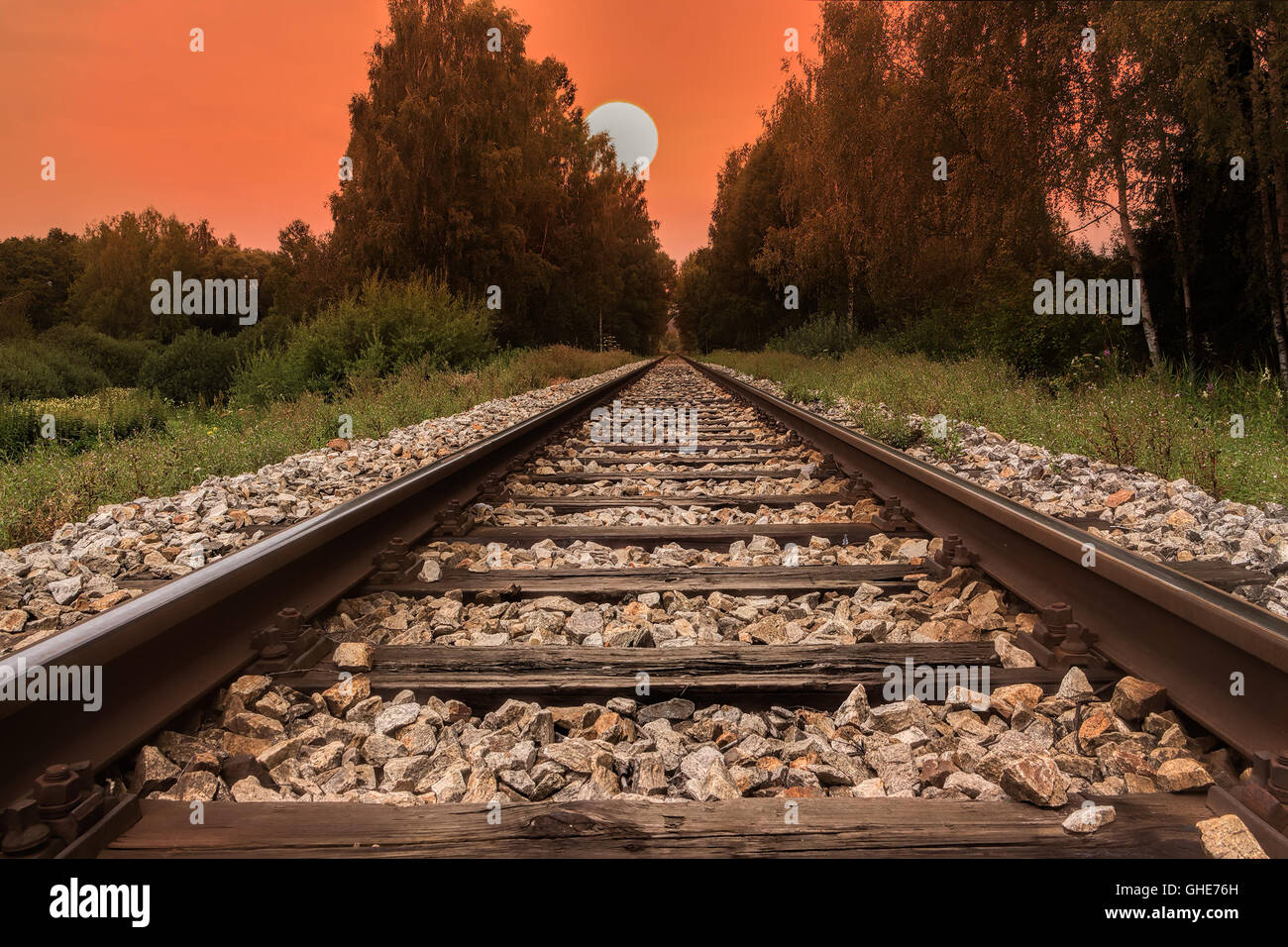 Railway line, trail or track  going through landscape at sunset or dawn. Red sky with big evening sun. Stock Photo