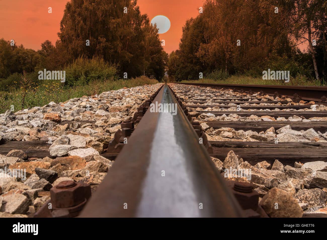 Railway line, trail or track going through landscape at sunset or dawn. Red sky with big evening sun. Stock Photo