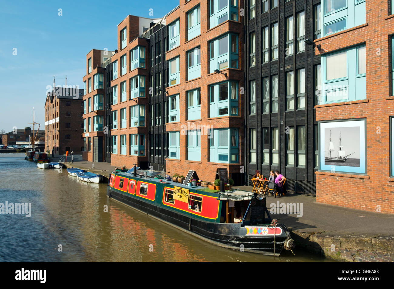 Canal boat cafe moored alongside new build apartments and shops in spring sunshine at Gloucester Docks, Gloucester, UK Stock Photo