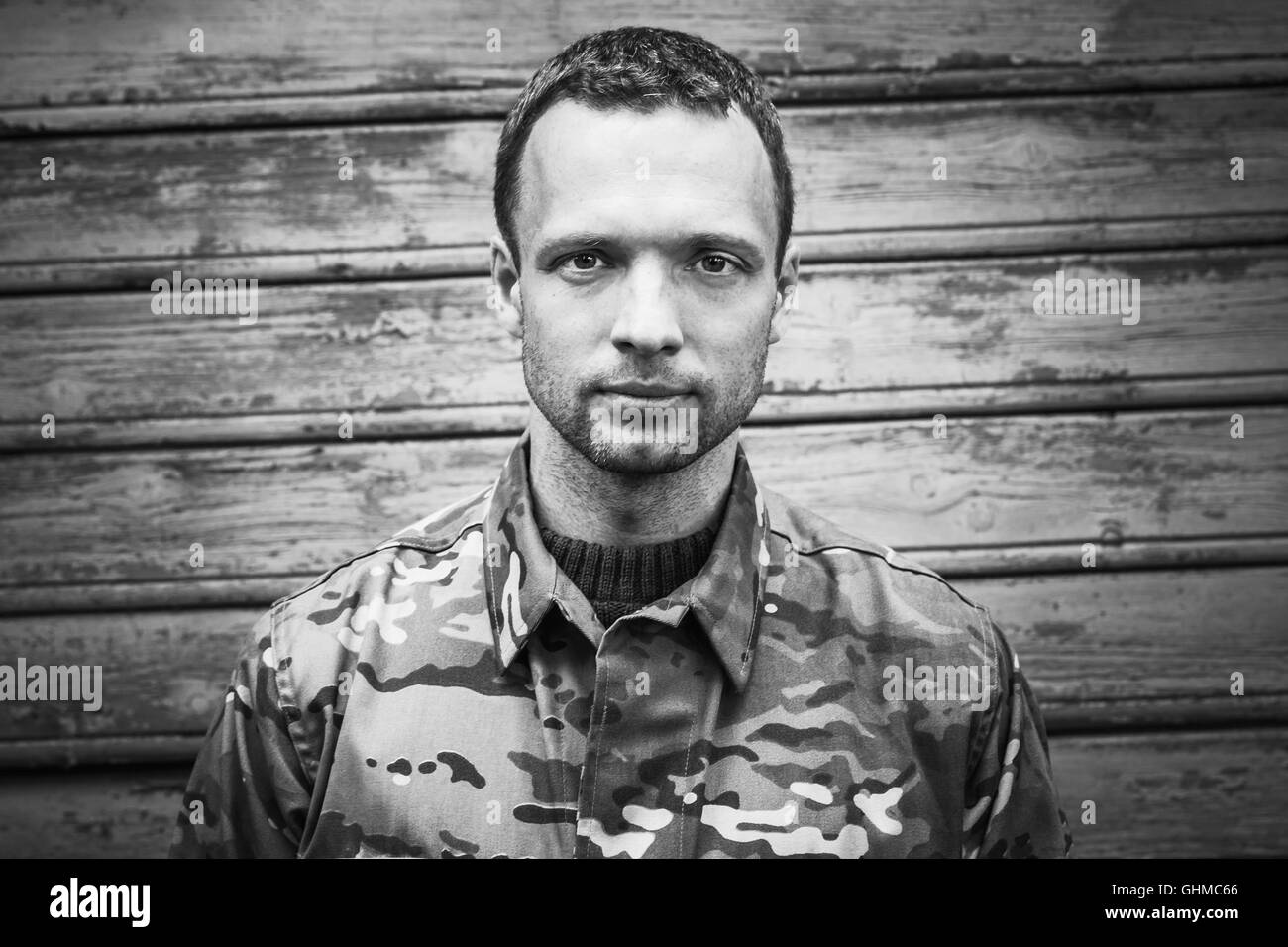 Young Caucasian military man in camouflage uniform. Closeup black and white frontal portrait over wooden wall Stock Photo
